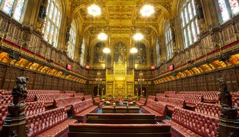 The House of Commons chamber, featuring rows of seats and a central podium, is depicted in a formal legislative setting.