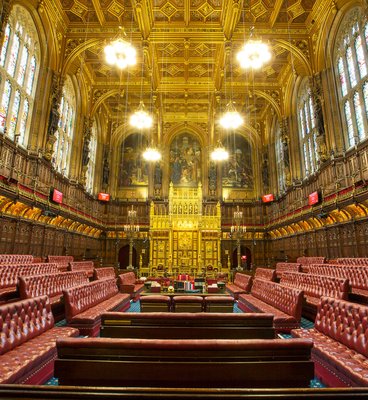 The House of Commons chamber, featuring rows of seats and a central podium, is depicted in a formal legislative setting.