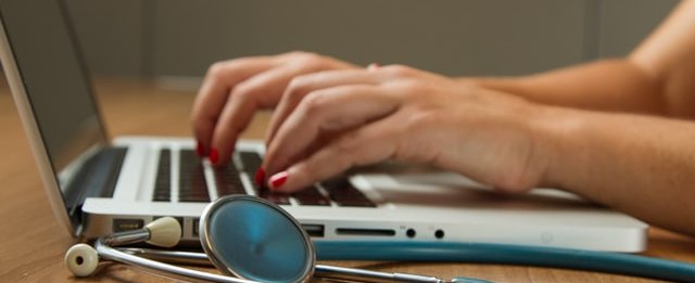 person typing on laptop - stethoscope next to laptop