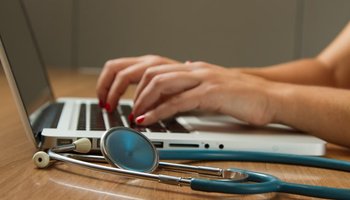 person typing on laptop - stethoscope next to laptop